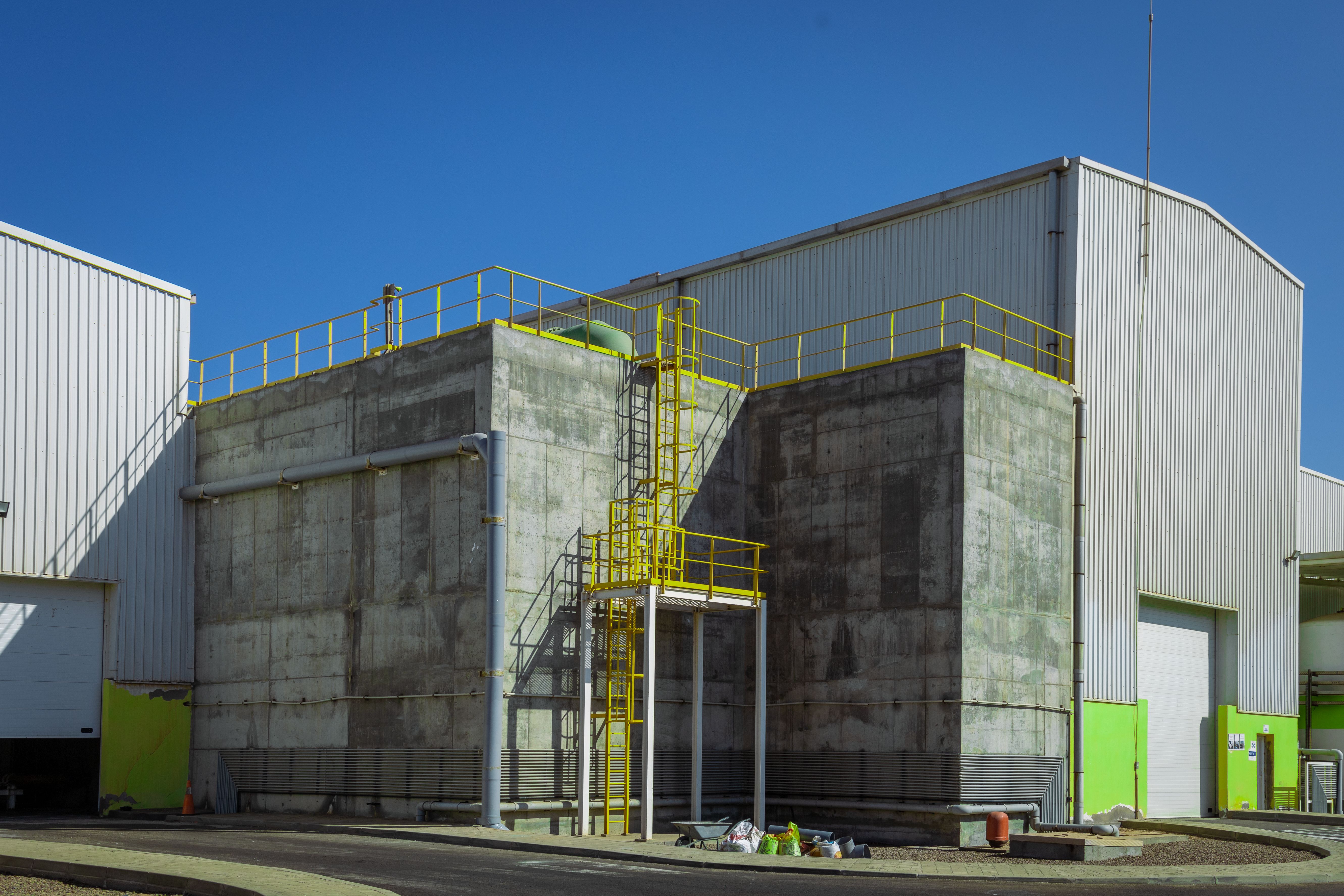 Fachada de galpão industrial moderno, com parede de concreto aparente, guarda-corpo amarelo e céu azul.